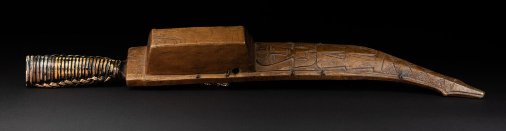 Short sword from the Paiwan tribe, with a sheath carved with a standing figure. The side of the sheath is adorned with three sculpted human heads. The sword is made in one piece with the handle, which is wrapped in rattan.