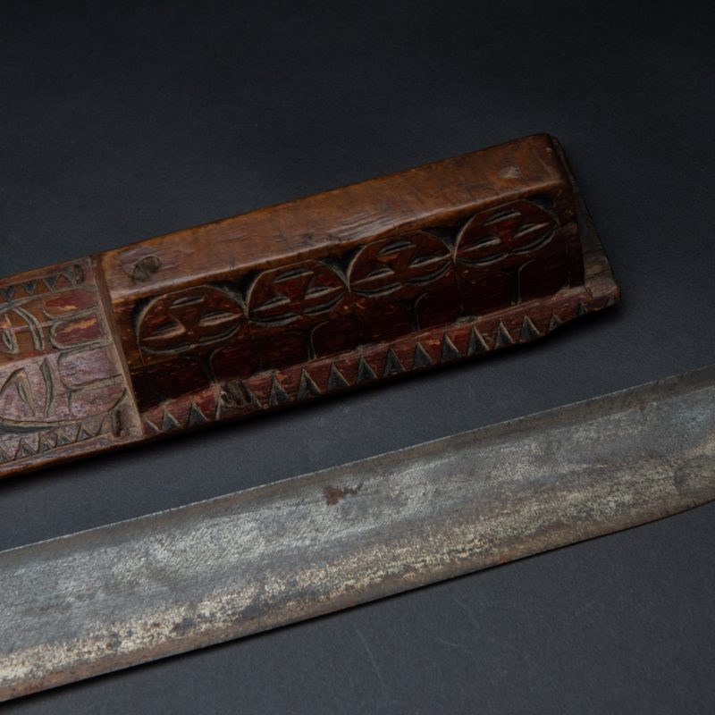 Paiwan tribe sword with a sheath carved with a standing figure and human faces on the side, peint in red. The hilt is made of carved wood.