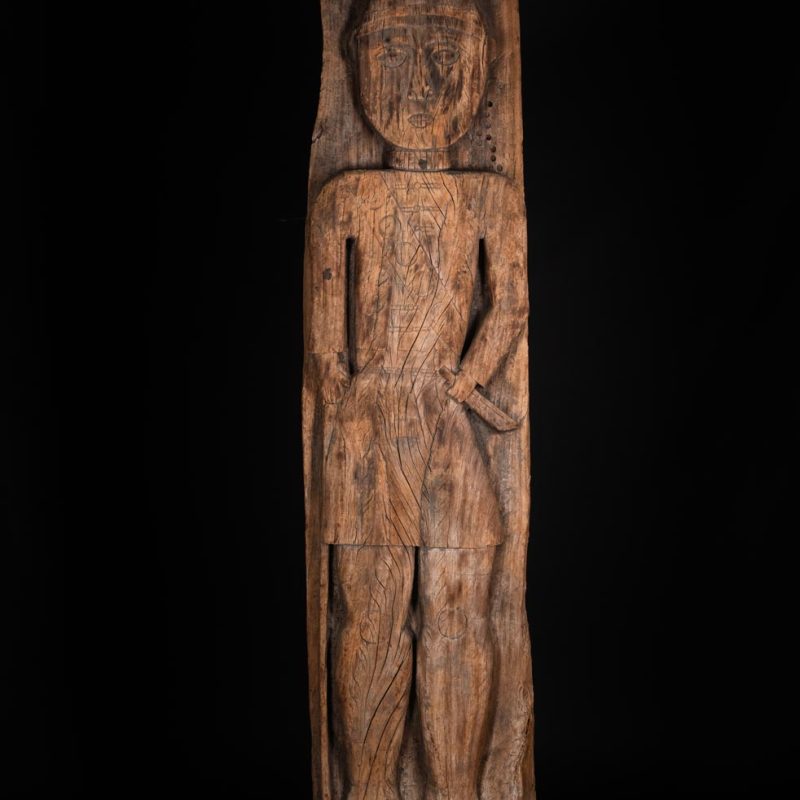 Paiwan tribe: massive 19th-century ancestral panel measuring over 2.5 meters, depicting a hunter wearing a Paiwan-style hat and holding a knife and a spear at his side. The carvings on his torso suggest he is dressed in a ceremonial vest and the shoulder belt traditionally worn by nobles. Due to its size, this panel was likely the central piece of the house.