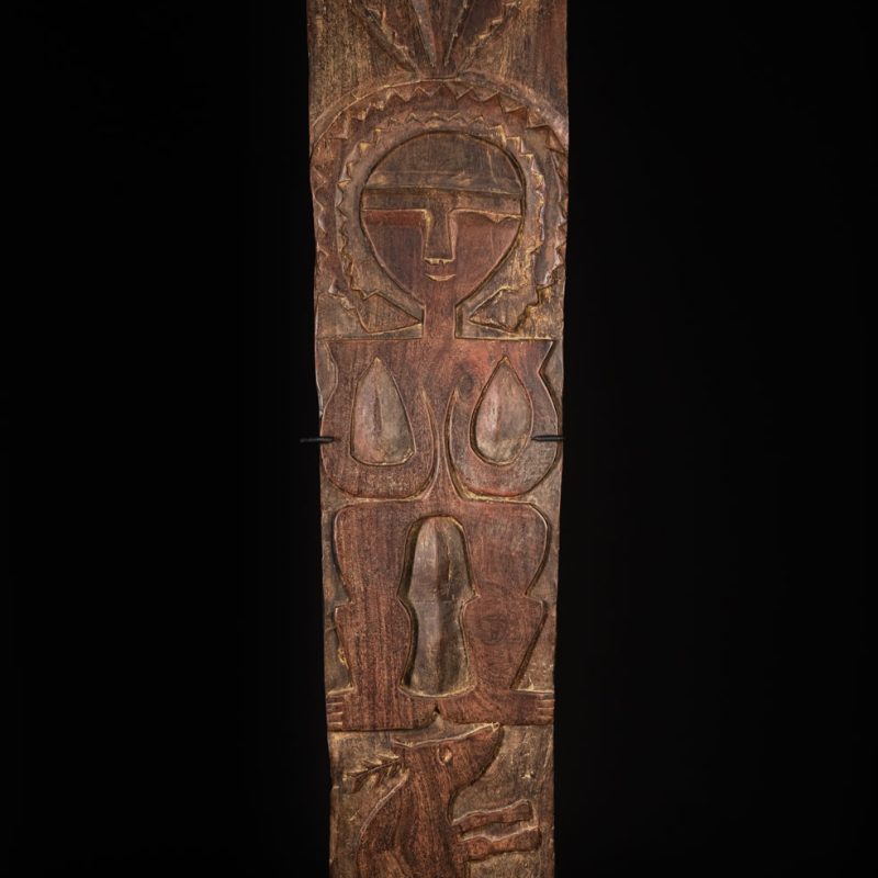Ancient panel from the Paiwan tribe, carved with a stylized ancestor wearing a two-headed snake headdress — a symbol of noble lineage — and eagle feathers, representing strength in battle. A deer lies at his feet.