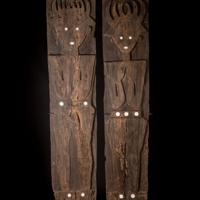 Pair of ancestor panels from the Paiwan tribe, depicting a male and a female figure, both wearing feathered headdresses — symbols of valor and vigor. The panels are inlaid with porcelain circles at the eyes, mouth, and waist, representing the ceremonial belts they wear.