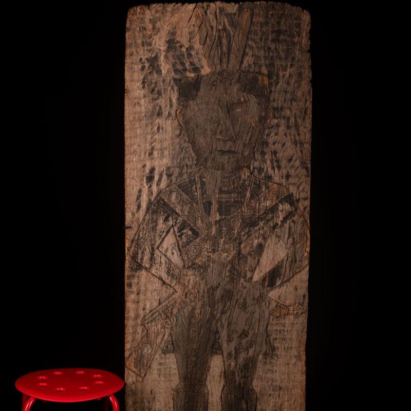 Old ancestor panel from the Paiwan tribe, carved with a standing warrior, one hand resting on his rifle and the other on the pommel of his sword. He is dressed in ceremonial attire and wears a hat adorned with eagle feathers, a symbol of valor.