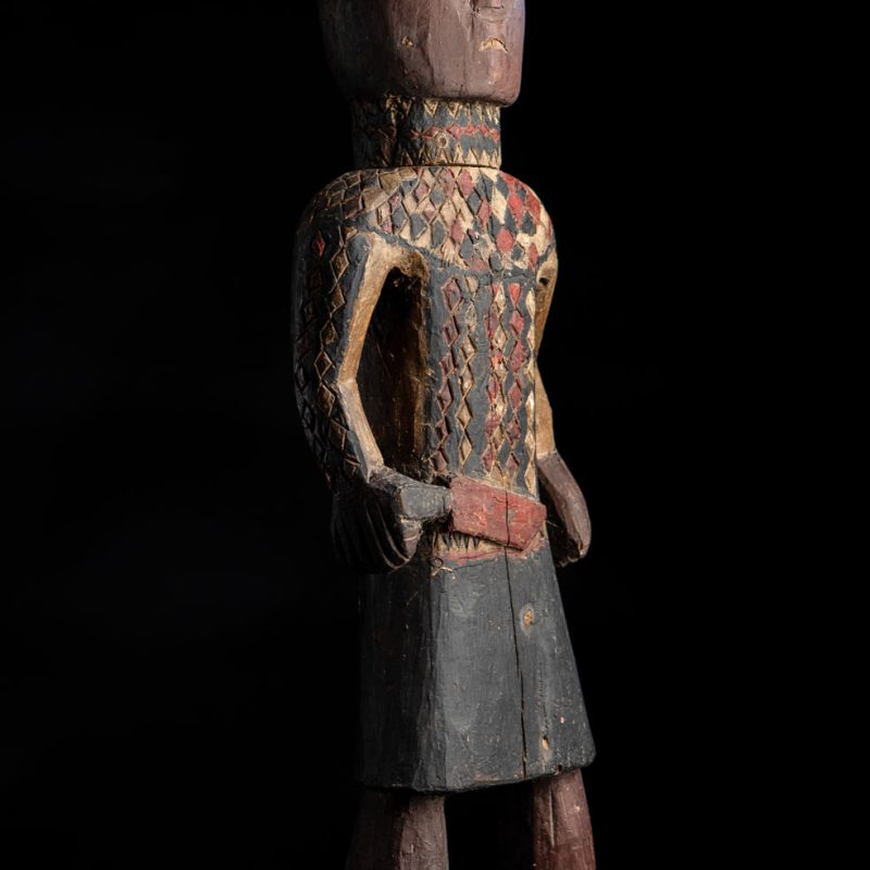 Wooden Statue from the Paiwan tribe painted in red, beige, and black, depicting a full-standing ancestor wearing ceremonial clothing and a leather headdress, with a knife at the waist.